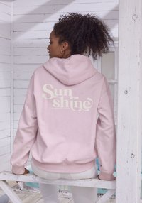 Felpa rosa con cappuccio e cordino; presenta la scritta bianca "Sun shine" sul retro. Tessuto morbido, polsi e orlo a costine, vestibilità comoda.