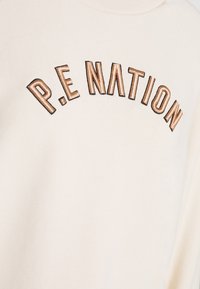 Ljus beige sweatshirt med "P.E NATION" broderat i mörkbrunt och metalliskt brons, med ribbad halsringning och mjuk textur.