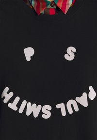 Sudadera negra con letras blancas de forma irregular que escriben "PAUL SMITH" dispuestas en una línea curva; camisa con colores llamativos debajo.