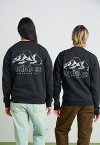 Melnā krekla ar kalnu grafiku un tekstu "adidas Originals"; brīva piegriezuma; valkāta kopā ar gaiši zilām un rakstainām brūnām biksēm.