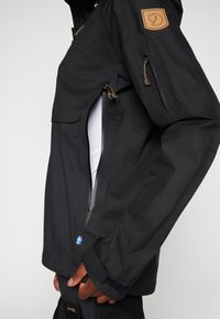 Veste imperméable noire avec une poche zippée, dotée d'un tissu texturé, de poignets élastiques et d'une étiquette en cuir beige sur l'épaule.