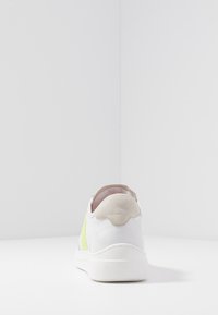 Baskets blanches avec un panneau latéral en mesh jaune et un accent talon en suède gris. Elles présentent une semelle en caoutchouc texturée et une tige en cuir lisse.