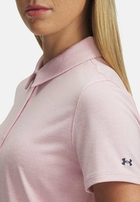 Ljusrosa polo-shirt med krage, korta ärmar och en slät yta. Har en liten marinblå logotyp på vänster ärm.