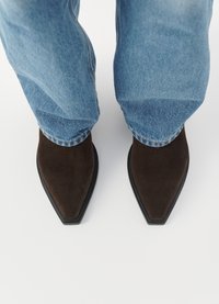 Stivali ankle in suede marrone con punta affusolata, caratterizzati da una suola nera piatta, indossati con jeans azzurri risvoltati.