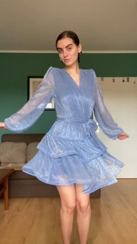 Απαλή γαλάζια φόρεμα με στάδια, μακριά διάφανα μανίκια, V-λαιμόκοψη και ζώνη στη μέση. Το ύφασμα έχει μια διακριτική λάμψη και μαλακή υφή.
