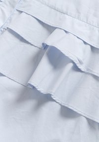 Lichtblauwe stof met gelaagde ruffles. Soepele textuur en zichtbare stiksels langs de randen en naden. Geen patronen of hardware aanwezig.