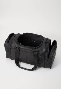 Sac de voyage en cuir noir avec une surface texturée, deux courtes poignées, un compartiment principal zippé et des poches zippées sur les côtés. L'intérieur est doublé en tissu.