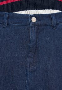 Σκούρο μπλε τζιν με κλείσιμο με κουμπιά, θηλιές για ζώνη και απαλή υφή. Ορατά ράμματα και ελαφριά λάμψη στο ύφασμα.