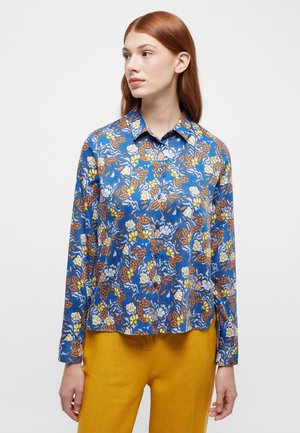 Blaues, blumiges Langarmshirt mit Kragen zum Knöpfen, verziert mit gelben und orangefarbenen Blättern und Blumen, präsentiert über einer senfgelben Hose.