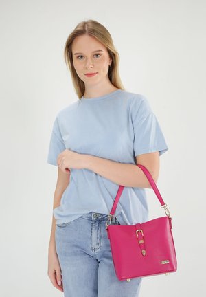Rosa Lederhandtasche mit strukturierter Form, ausgestattet mit einer goldenen Hardware-Schnalle und einem abnehmbaren Schultergurt. Das Model trägt ein hellblaues T-Shirt.