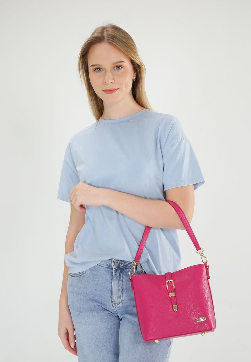 Roze leren handtas met een gestructureerde vorm, voorzien van een gouden hardware gesp en een afneembare schouderband. Model draagt een lichtblauw T-shirt.