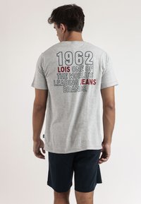 Camiseta de algodón gris con un gran texto en la parte posterior: "1962 LOIS UNA DE LAS MARCAS DE VAQUEROS MÁS IMPORTANTES DEL MUNDO." Diseño básico de cuello redondo.