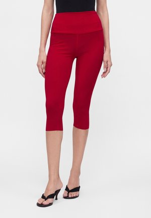 Femme portant des leggings capri rouges, moulants et taille haute, ainsi que des sandales noires à talons ouverts, debout devant un fond uni.