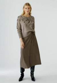 Beiger Pullover mit floralen Stickdetails, kombiniert mit einem langen, olivgrünen Wickelrock. Das Model trägt elegante, schwarze kniehohe Stiefel.