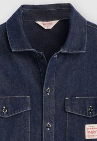 Chemise en denim foncé à coupe décontractée, boutonnée sur le devant, avec deux poches poitrine et des coutures contrastantes blanches. L'étiquette indique « travail à faire ».