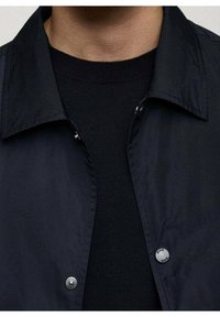 Veste légère bleu marine au design élégant, dotée d'une fermeture à boutons, d'un col pointu et d'une texture lisse. Pull noir visible en dessous.