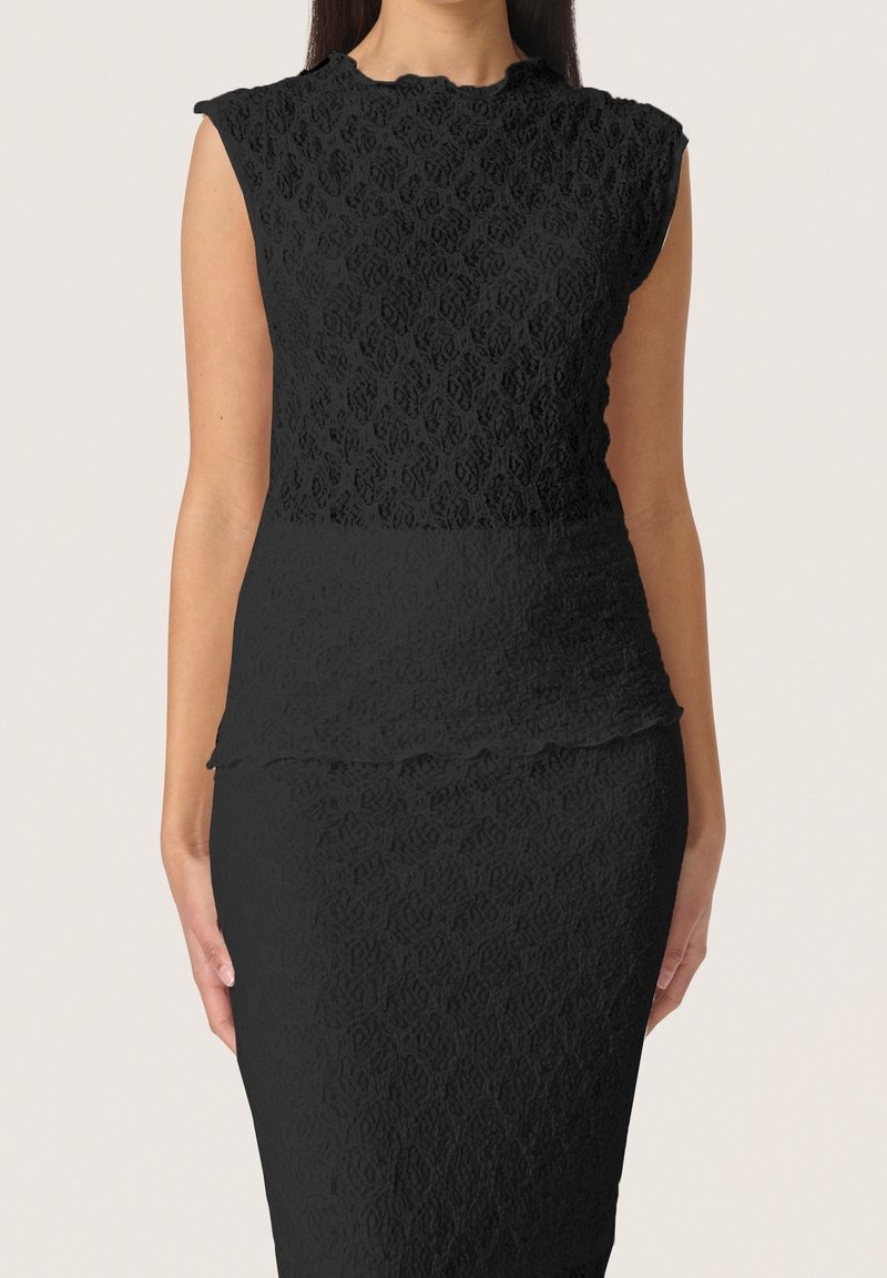 Femme portant une robe noire en dentelle sans manches, avec une taille cintrée et un léger détail péplum sur un fond uni.