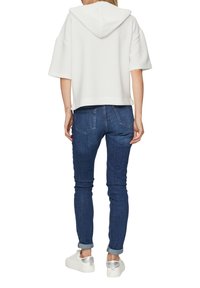 Sweatshirt à capuche blanche avec un motif texturé, associé à un jean skinny bleu foncé. Le modèle porte des chaussures blanches avec un accent argenté.