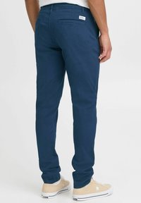 Pantalones slim-fit azul marino de algodón con dos bolsillos traseros y una pequeña etiqueta de marca en la cintura. Llevados con zapatillas beige de corte bajo.