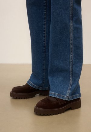 Jambes portant un jean bleu large et des mocassins marron foncé à semelle épaisse, debout sur un sol beige.