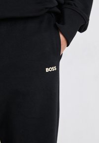 Melni sporta bikses, izgatavotas no mīkstas auduma, ar niansētu zelta "BOSS" logotipu uz augšstilba un sānu kabatu.