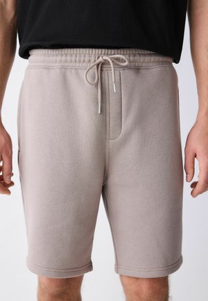 Mand iført beige snøre-shorts og en sort skjorte, der viser fronten af talje og øvre benområde mod en ensfarvet baggrund.