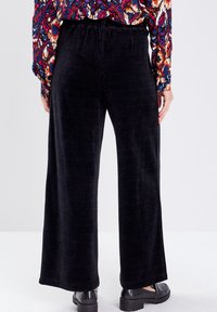 Pantalons larges en velours côtelé noir avec une texture à côtes verticales, dotés d'une taille élastique et d'une coupe décontractée, associés à des chaussures noires.