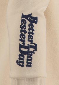 Manga de suéter beige con texto azul oscuro que dice "Mejor que Ayer" en una fuente audaz y estilizada. La tela parece suave y texturizada.