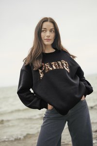 Crna majica s natpisom "PARIS" u dizajnu životinjske print, okrugli izrez, širok kroj, uparena s tamno sivim trapericama. U pozadini se vidi voda i oblačno nebo.