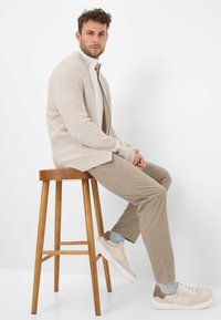 Beige gebreide cardigan met hoge kraag, gecombineerd met lichtgekleurde, gestructureerde broek en beige sneakers. Model zittend op een houten kruk.