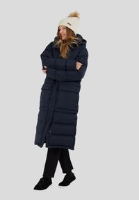 Námořnický modrý péřový dlouhý kabát s kapucí, se dvěma předními kapsami a elegantním prošívaným designem. Nesený s krémovým pleteným čepichem.
