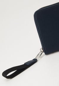 Portafoglio blu navy con finitura testurizzata, dotato di una zip silver e di un cinturino da polso in tessuto nero per una facile maneggevolezza.