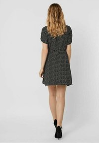 Schwarzes Kleid mit weißen Pünktchen, kurzen Puffärmeln, tailliertem Schnitt, ausgestelltem Rock und einem Saum, der bis zur Mitte des Oberschenkels reicht; getragen mit schwarzen Absatzschuhen.