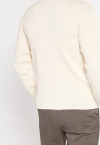 Sweter dziany w kolorze kremowym z okrągłym dekoltem i prążkowaną fakturą. Noszony z brązowymi spodniami o fakturze, prezentujący tylny widok i detale rękawów.