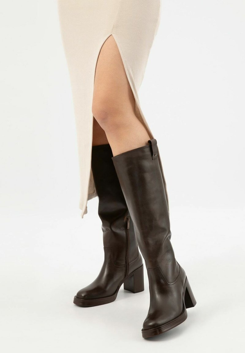 Bottes en cuir marron jusqu'aux genoux avec un talon carré, dotées d'une fermeture éclair sur le côté et d'une texture lisse, associées à une robe crème avec des fentes.