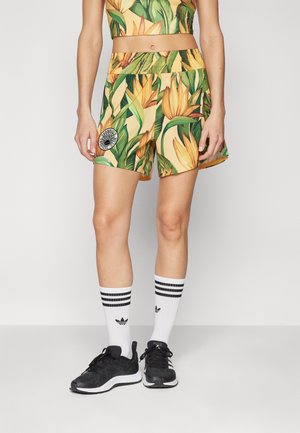 Ben iført shorts med tropisk print og matchende top, hvide Adidas sokker med sorte striber og sorte løbesko på en ensfarvet baggrund.