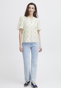 Gul blus med blommönster, puffärmar, v-ringning och texturerad tyg. Matchad med ljusblå utsvängda jeans och blå sneakers.