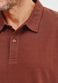 Brązowa bawełniana koszulka polo z luźnym kołnierzykiem, dwoma guzikami i teksturowanym splotem, podkreślająca finezyjne detale szycia.