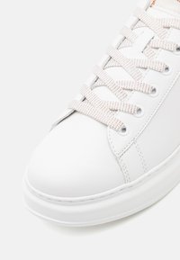 Shoe branco com parte superior sintética texturizada, bico redondo e sola de borracha plana. Apresenta atacadores brancos com sutis acentos laranja.