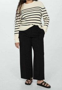 Pull rayé crème et noir à manches longues associé à un pantalon noir ample ; le modèle porte des sandales noires à lanières et pose les mains dans les poches.