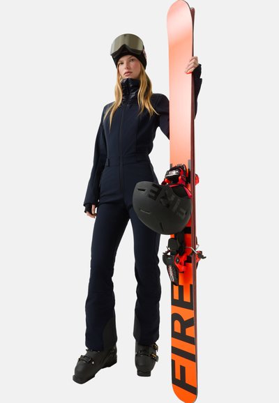 Tmavě modrý lyžařský overal s vysokým límcem, spojený s oranžovým snowboardem s černými detaily a černou helmou, držený osobou.