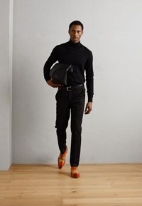 Maglione nero a collo alto, pantaloni neri, scarpe di pelle arancioni. Modello con borsa nera, in piedi su un pavimento in legno contro una parete chiara.