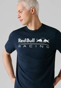 Junger Mann mit kurzen blonden Haaren, der ein marineblaues Red Bull Racing T-Shirt trägt, schaut nach links und hat die Hände in die Hüften gestemmt vor einem grauen Hintergrund.