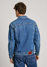 Chaqueta de mezclilla con un collar clásico, mangas largas y un teñido azul vibrante. Incluye una etiqueta de cuero marrón en la parte trasera de la cintura y costuras visibles.