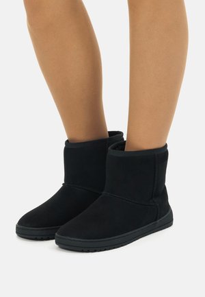 Botas de tobillo de ante negro con una textura suave, con puntera redonda y detalles de costura, montadas sobre una suela de goma plana.