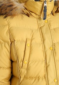 Gelbe Pufferjacke mit einem mit Fell gefütterten Kapuze, Reißverschluss vorne, zwei Klappentaschen und sichtbaren Nähten auf einer gesteppten Textur.