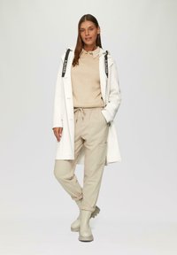 Cappotto lungo bianco con dettagli neri, felpa beige e jogger beige con tasche. Stivali in gomma beige completano l'outfit.