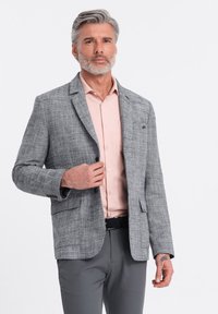 Grå texturerad kavaj med tre knappar, framfickor, bärs över en ljusrosa skjorta. Grå byxor och ett svart bälte fulländar outfiten.