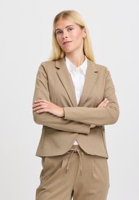 Beige kohandatud bleiser pehmest ja tekstuurist kangast, millel on nurgalapid ja avatud esiosa, koos sobivate beežide pükstega.