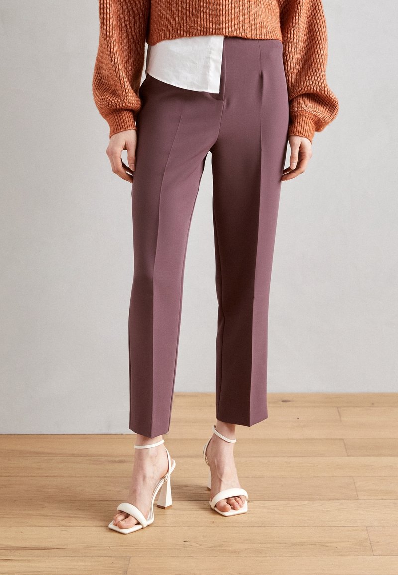 Femme debout sur un sol en bois portant un pantalon mauve ajusté, une chemise blanche, un pull rouille et des sandales blanches à talons hauts. Buste coupé.